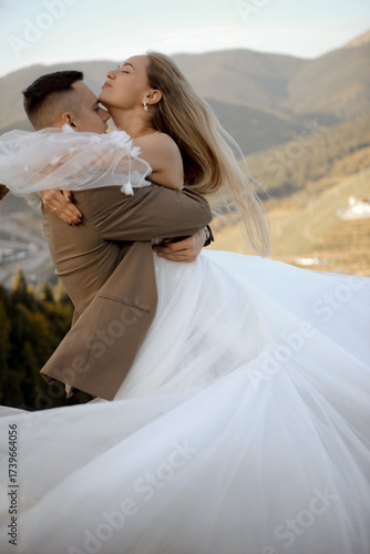A beautiful, happy young couple in love on a wedding walk and photo shoot high in the mountains. The groom in a jacket and suit and the bride in a white dress at a resort. Kissing and hugging