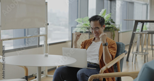 Senior Asian businessman wearing headset celebrates success during online meeting in modern co-working space. Remote work and business communication concept	