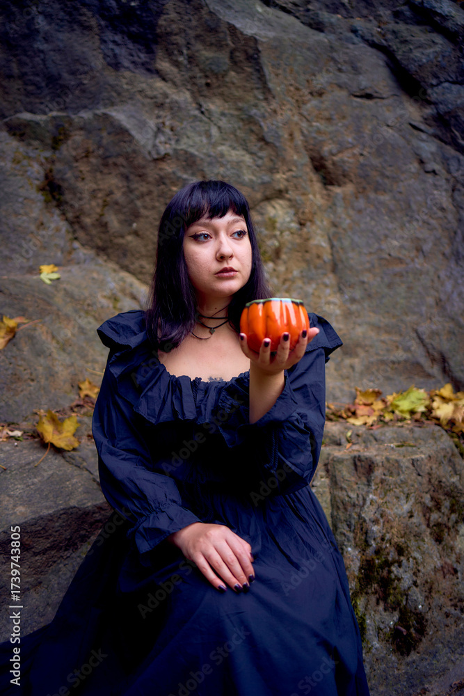 Obraz premium woman in a black long dress sits on black rocks on Halloween holding a pumpkin-shaped cup, a modern witch