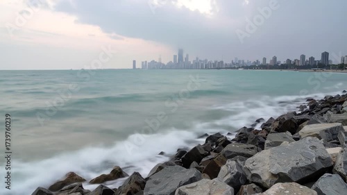 Long exposure of waves crashing on a rocky shore with a distant city skyline under a cloudy sky