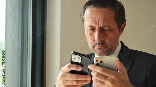 Confused businessman using two smartphones simultaneously, trying to synchronize data between devices. 4K.
