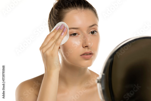 Studio shot of young woman with no makeup cleaning face with cotton pad in mirror on white background.