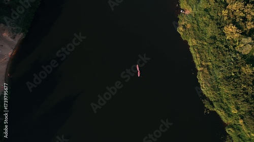 Two young women in life jackets lie on pink SUP boards in black water of river surrounded by picturesque banks. View from above.