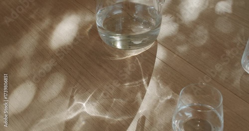 Sunlight streams through a window, casting dancing shadows on a wooden table where a glass pitcher of water sits beside two clear glasses. One glass is empty, the other half full,