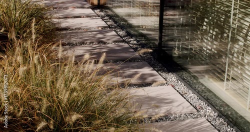Modern Pathway by Glass Wall. A serene walkway with stepping stones, ornamental grass and gravel. Sunlight filters through nearby trees, casting shadows on the path and glass facade. architecture
