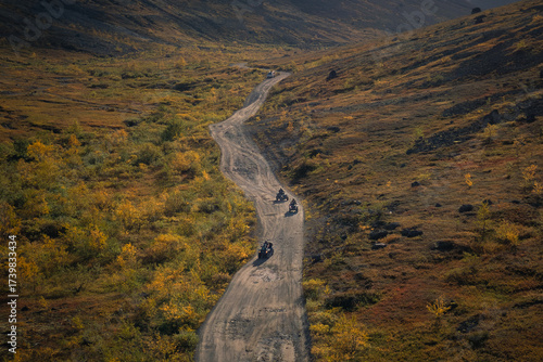 ATV riders traveling along a winding dirt road in the autumn tundra, with mountains in the background. Travel in Lapland