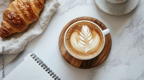 Close-up of a coffee cup with latte art on a wooden saucer alongside a croissant and a notebook on marble surface in a cozy cafe setting