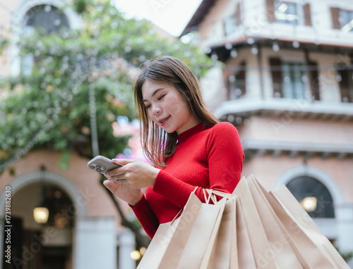 A young woman in a red sweater holding multiple shopping bags and using her smartphone while smiling in a modern shopping area.