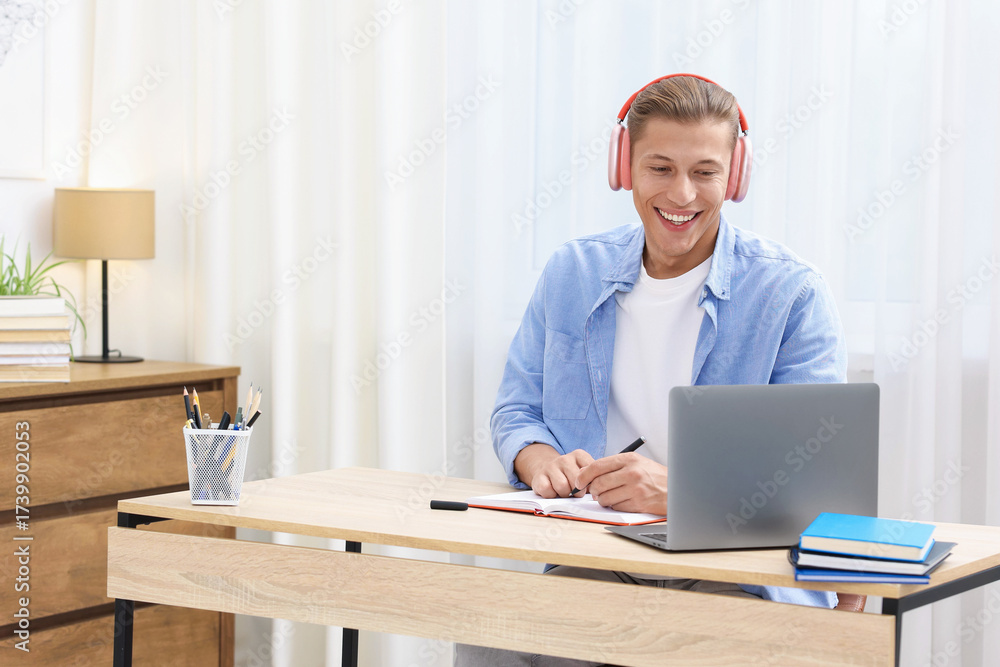 Fototapeta premium Man in headphones taking notes while passing online test via laptop at table indoors