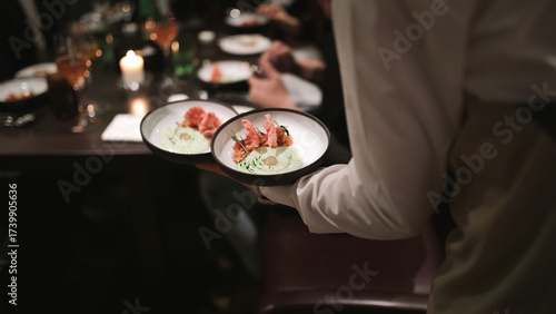 Waiter bringing out two bowls of a crudo dish on one hand during busy seafood night at the restaurant.