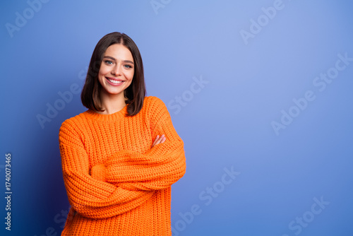 Smiling young woman in orange knitted sweater posing confidently against a purple background, exuding cheerful and stylish vibes