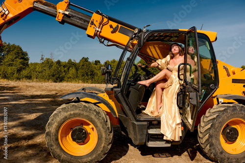 Elegant woman in a flowing golden gown playfully relaxes inside a yellow telescopic handler against the backdrop of a sunny construction site, creating an intriguing blend of fashion and industry.