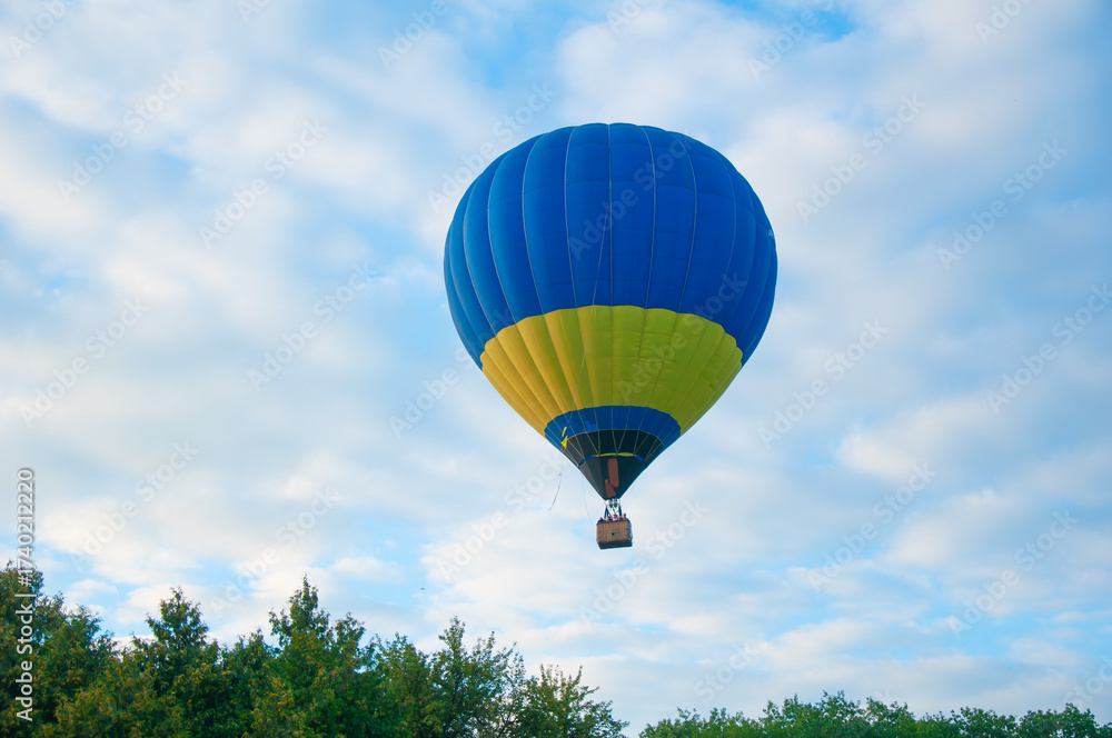 Fototapeta premium Blue and yellow hot air balloon floating above green trees under a partly cloudy sky. A serene aerial scene blending adventure and national colors