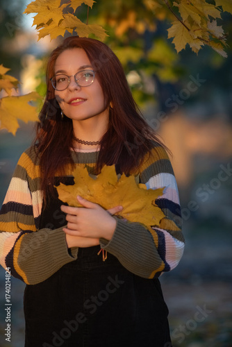 Young woman with glasses, holding vibrant autumn leaves, stands in a sunlit outdoor setting, surrounded by colorful foliage, capturing the essence of fall and nature's beauty