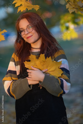 Young woman with glasses, holding vibrant autumn leaves, stands in a sunlit outdoor setting, surrounded by colorful foliage, capturing the essence of fall and nature's beauty