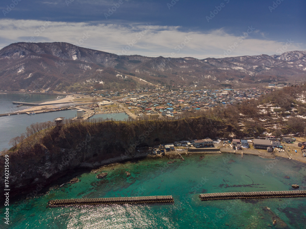 Naklejka premium Aerial view of a coastal town with turquoise waters, harbor structures, and snow-dusted mountains in the background