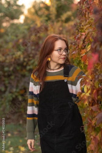 Young woman with glasses, holding vibrant autumn leaves, stands in a sunlit outdoor setting, surrounded by colorful foliage, capturing the essence of fall and nature's beauty