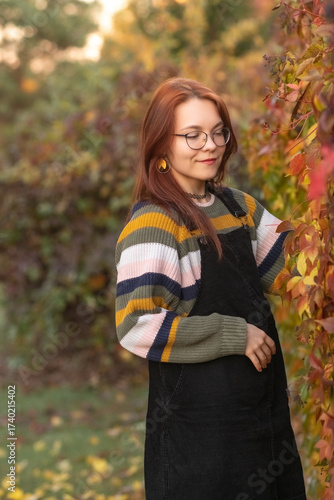 Young woman with glasses, holding vibrant autumn leaves, stands in a sunlit outdoor setting, surrounded by colorful foliage, capturing the essence of fall and nature's beauty