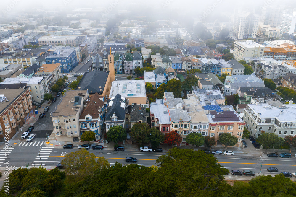 Obraz premium Aerial view of colorful Victorian style houses in San Francisco with a tree lined street, parked cars, and a church steeple in the foggy background.
