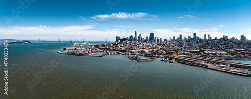 Aerial view of San Francisco, California, showing the downtown skyline, Bay Bridge, Alcatraz Island, waterfront piers, and docked boats under a clear sky.