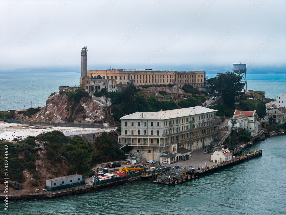 Obraz premium Alcatraz Island in San Francisco Bay features the iconic prison, lighthouse, water tower, rugged cliffs, greenery, dock area, and surrounding waters.
