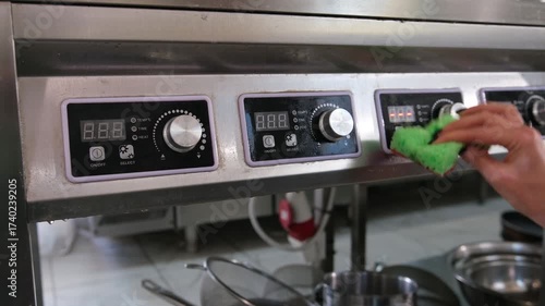 Restaurant worker's hand carefully wiping a stainless steel industrial stove with a green sponge, ensuring cleanliness and hygiene standards in a professional commercial kitchen environment
