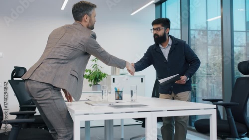 Caucasian man in suit shaking hands with South Asian man during job interview. Confident gesture at meeting in modern office. CV or resume reading. Interviewing for new work position in company.