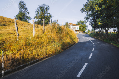 Curved rural road leading uphill through golden autumn grass and trees, with a small house at the top under clear blue sky, peaceful countryside landscape