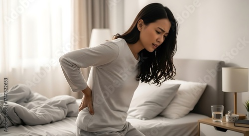 Young woman suffering from back pain while sitting on bed in morning
