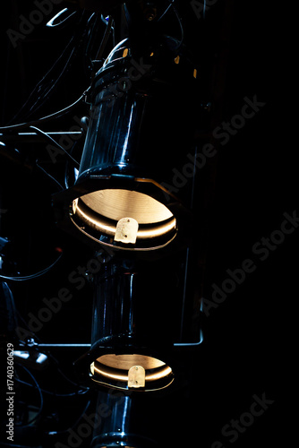 Two theatrical spotlights with warm, golden illumination hang vertically in absolute darkness. The scene is dramatic and moody, highlighting professional stage equipment.

