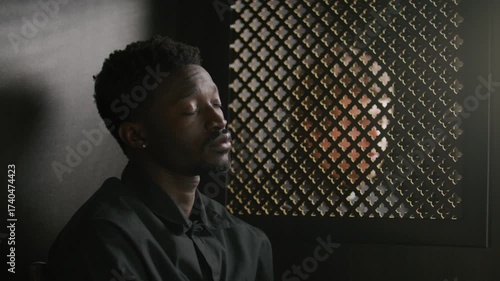 Young African American man sitting in confessional booth with ornate partition screen, answering questions from priest during confession