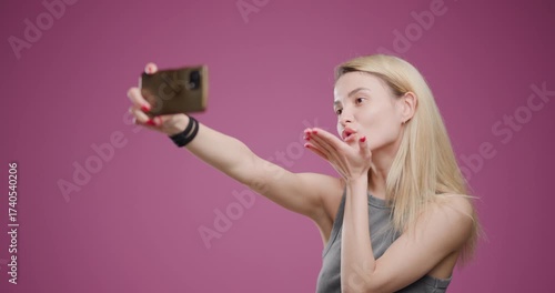 Young blonde woman taking a playful selfie, puckering lips on a pink background