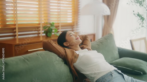 Relaxed young Asian woman enjoying rest on comfortable sofa, calm attractive girl relaxing on couch, breathing fresh air with eyes closed, meditating at home