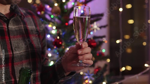 Caucasian man clinking bottle with champagne glass on holiday, against backdrop of garlands and Christmas tree close up. Lonely unrecognizable man Christmas celebration.