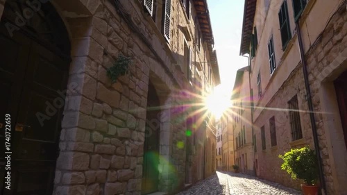  Slow forward movement shot in a narrow cobblestone alley of an old European town with a bright sun flare appearing between the historic stone buildings.