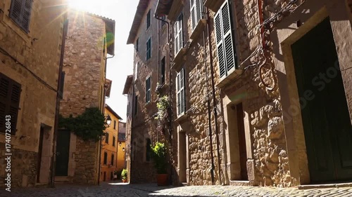 Static shot of a narrow cobblestone street in a historic European town with old stone buildings and a bright sun flare.