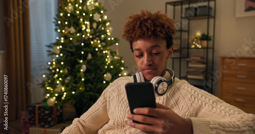 Boy in winter warm sweater relaxes on couch spends time alone with wireless headphones listening to podcast, changes songs on phone, laughs, Christmas tree in background.