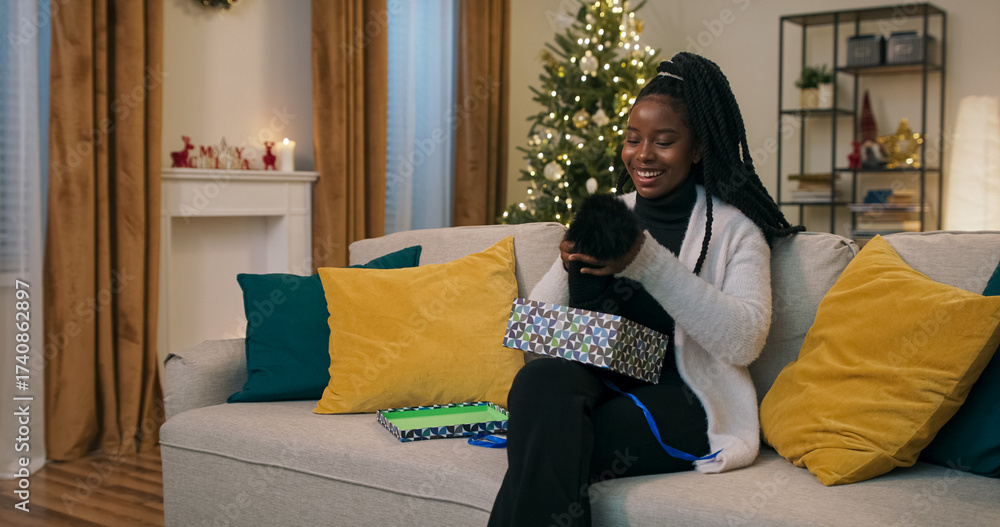 Obraz premium A young student of multi-ethnic background sits on a couch, unpacking a Christmas present. Expecting a miracle, she opens the box, pulls out a warm black hat, rejoices, and tries it on.