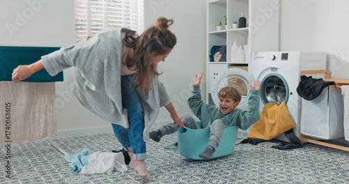 A cheerful son plays in the laundry room, sitting inside a bowl of clothes. His mother gently pulls him, enjoying their playful moment during chores.