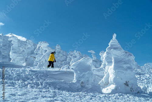 A traveler in a yellow jacket walks through snow-covered trees, known as snow monsters, under a clear blue winter sky at Mount Zao, Yamagata, Japan.