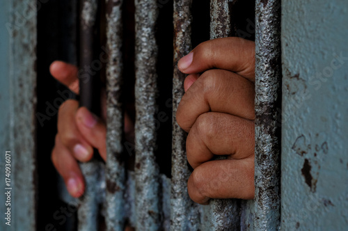 A person's hands tightly grasp the vertical, weathered metal bars of a rusty prison door or window, emphasizing feelings of confinement and despair in a stark, dimly lit setting.