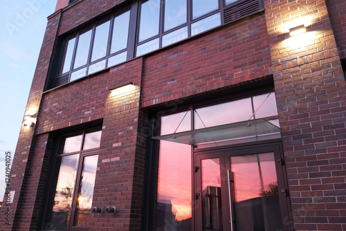 A burgundy brick wall of a residential building with windows and streetlights. The red sunset is reflected in the glass windows.