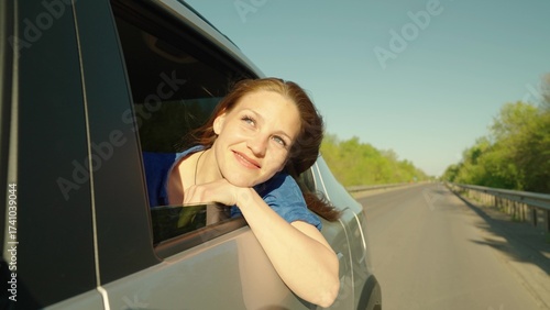Free girl travels smiling from car window, warm wind blowing her hair. Road trip on way to vacation. Young woman passenger looks out window, enjoys fresh breeze from car window, Happy look into future