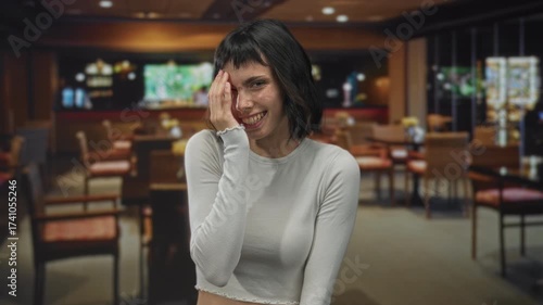 Young hispanic woman smiling broadly with hand covering face in a cozy restaurant building; embarrassment.