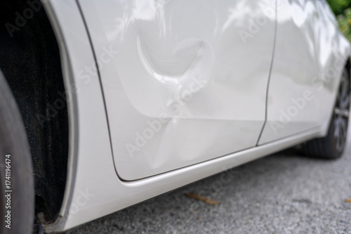 Close-up view of a dented car door on a white vehicle after an accident, showing the impact damage and body panel deformation on a residential street.