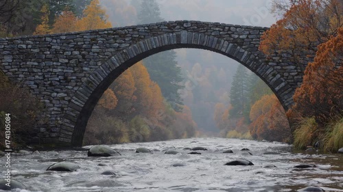 Old stone arch bridge spanning misty river surrounded by autumn mountain ash trees and forest hills