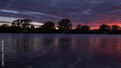 Fiery sunset sky reflected in calm lake beneath silhouettes of sycamore trees on distant shoreline