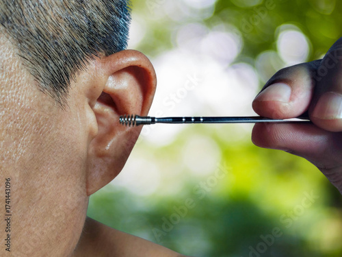 A man is putting ears pick metal cleaning his left ear for healthcare on a green blurred tree background.