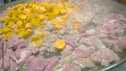 Chunks of raw chicken and sliced carrots frying together in a large pan with steam rising during cooking, showing the beginning stage of a stew or sauté dish preparation.