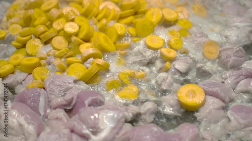 Close up shot of chunks of raw chicken and sliced carrots frying together in a large pan with steam rising during cooking, showing the beginning stage of a stew or sauté dish preparation.
